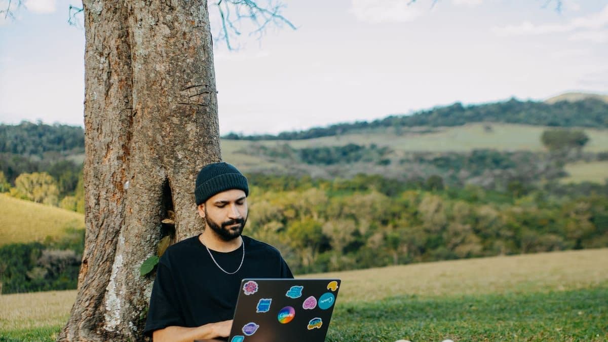 A freelancer working on a laptop under a tree in a green hilly landscape